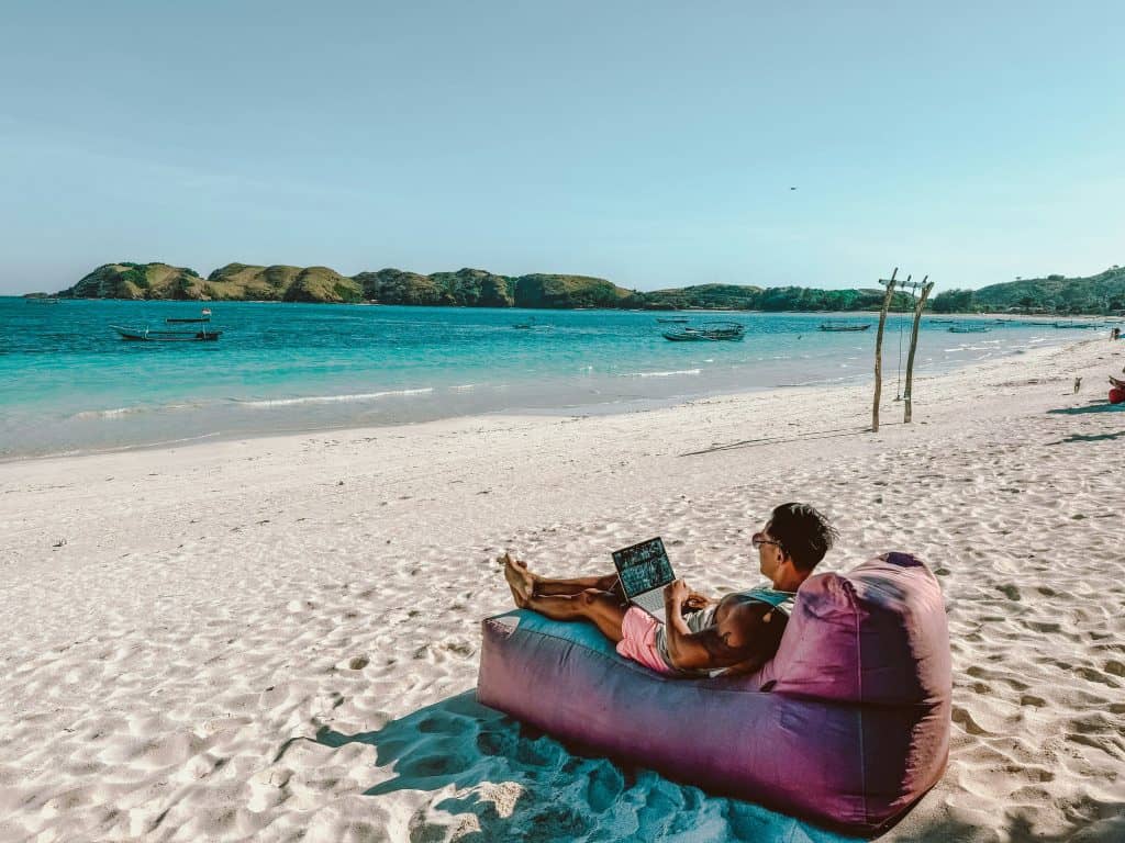 Man working on a laptop while sitting on a bean bag on a beach in Lombok, Indonesia, representing the concept of working remotely in another country​