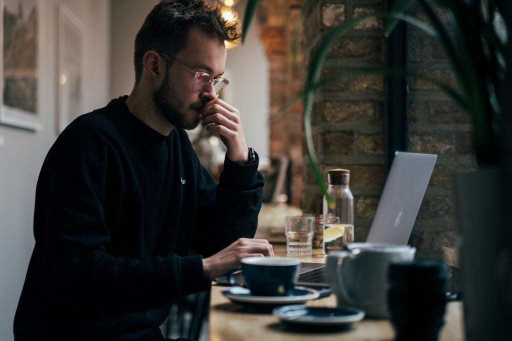 Man working on a laptop at a table with coffee cups and water glasses, representing the comparison between Bizee and LegalZoom services​