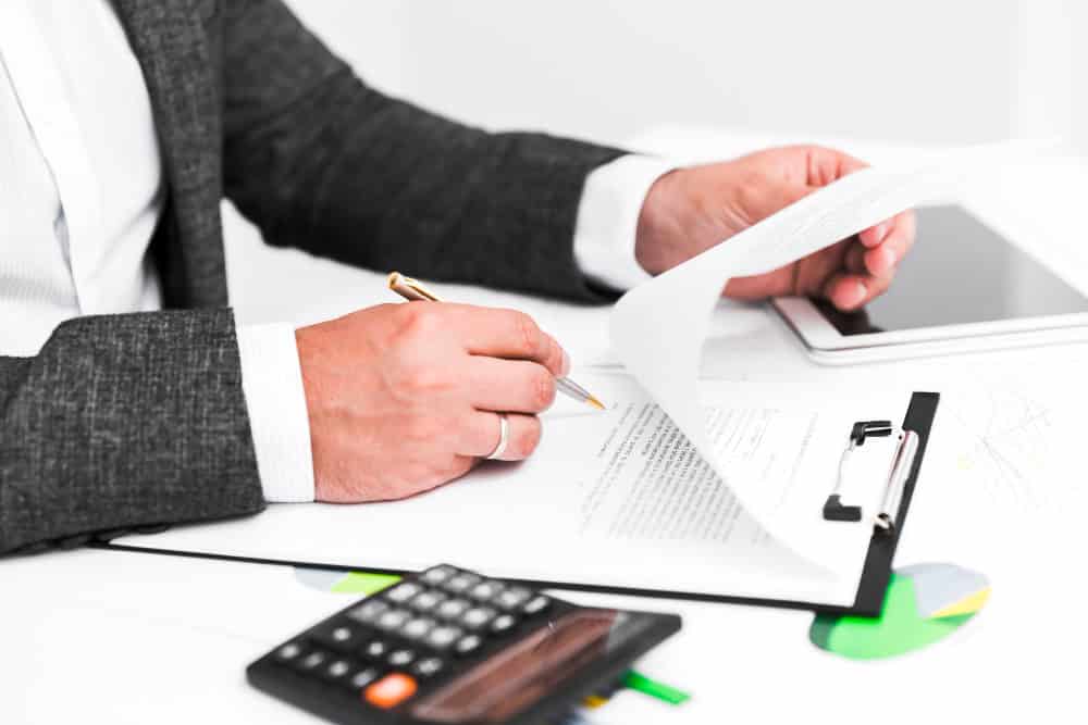 Detailed view of a professional hand reviewing business documents with charts, calculator, and tablet in an office setting. Focus on business paperwork, analysis, and professional work environment.