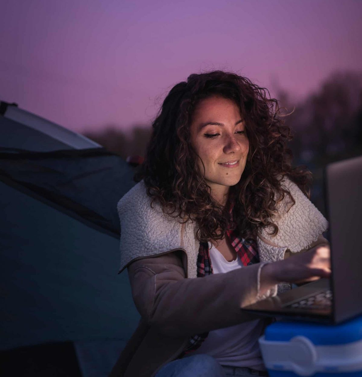Young woman camping by the lake, sitting on the tent entrance and working on a laptop computer;  digital nomad lifestyle